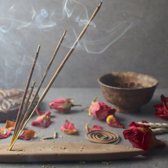 An image of lit incense burners over a wooden tray surrounded by picked up roses. There is smoke in the image